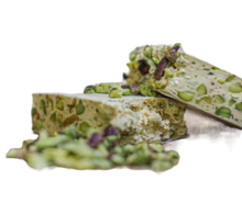 Lade das Bild in den Galerie-Viewer, Zwei Stücke Nove TORRONE MORBIDO AL PISTACCHIO 150g mit grünen Pistazien sind zwischen verstreuten Nüssen auf weißem Hintergrund gestapelt und betonen den Geschmack des sizilianischen Honigs bei jedem Bissen.