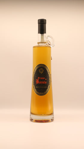 Eine hohe, klare Glasflasche Salvano GRAPPA DI BAROLO RISERVA 0,7 / 1,5 Liter mit bernsteinfarbener Spirituose im Inneren verfügt über ein schwarz-goldenes, verziertes Etikett und einen runden Glashals, der vor einem beigen Hintergrund steht.