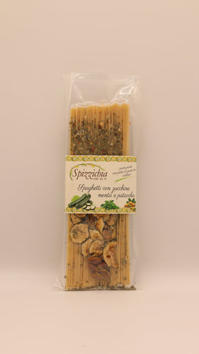 Eine durchsichtige Plastikverpackung mit Spizzicchia Spaghetti con zucchine, menta e pistacchio (200 g) mit getrockneten Zucchini, Minze und sizilianischen Pistazien. Auf dem Etikett steht der Produktname. Der Hintergrund ist einfarbig und hell.