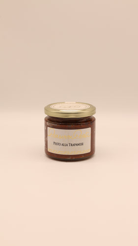 Ein Glas mit Conserve conti Pesto alla Trapanese - 190 g steht in der Mitte eines beigen Hintergrunds, auf dem Mandeln wie bei traditionellem sizilianischem Pesto aus Trapani abgebildet sind, mit einem goldenen Deckel und einem Produktetikett.